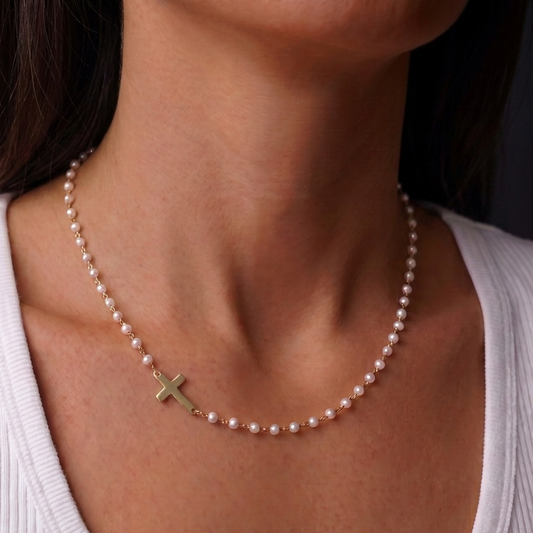 A close-up of a woman's neck and upper chest wearing a delicate pearl necklace with a small gold cross pendant, paired with a white ribbed tank top.